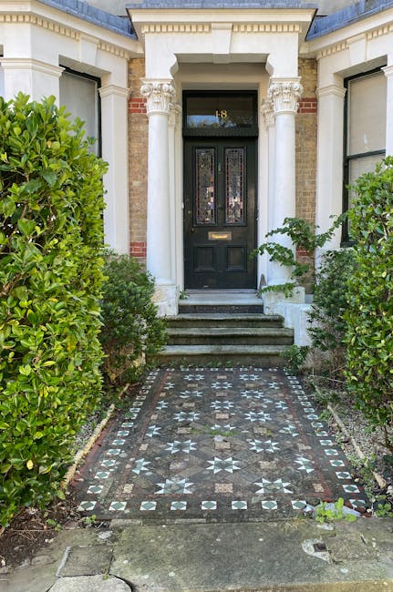 The image depicts the front entrance of a classical-style residential building with a black wooden door featuring decorative glass panels. The door is framed by two white painted columns with ornate capitals, supporting a small overhang. A brass mail slot is visible on the lower part of the door. Leading up to the entrance are three grey stone steps, with the bottom step partially obscured by a mosaic tiled pathway. The pathway is decorated with intricate geometric patterns in shades of grey, white, and black, and is bordered by small paving stones. Flanking the pathway are lush, neatly trimmed green bushes, which extend upward on both sides, creating a symmetrical framing of the entrance. The building's facade is constructed of cream-colored brickwork, with additional decorative brick and stone detailing near the window sills, which are partially visible on either side of the entrance. The overall scene is well-lit by natural daylight, emphasizing the contrast between the dark door and the lighter surrounding architectural elements, and it captures a tidy, well-maintained front garden and entrance area typical for private residential properties engaged in independent waste management or rubbish clearance services like those offered by Rubbish Clearance Holland Park.