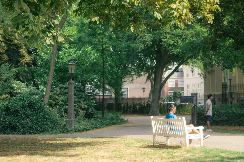 A park scene featuring a white wooden bench placed on a paved pathway, with a woman seated on it wearing a blue shirt and shorts, facing to the right. To the right of the bench, a young girl in a white top and dark shorts stands on the pathway, looking into the distance. Behind her, a large tree with green leaves and a sturdy trunk provides shade, and several smaller bushes and plants line the edges of the park. A classic black lamppost is visible near the left side of the image, surrounded by lush greenery, including various shrubs and other trees with mixed green foliage. In the background, residential buildings with windows and balconies are partially visible through the dense trees and park fencing. The scene is well-lit by natural daylight, indicating a clear day, and the overall environment appears to be a peaceful urban park suitable for relaxation and casual outdoor activities, fitting within a typical residential neighbourhood setting.
