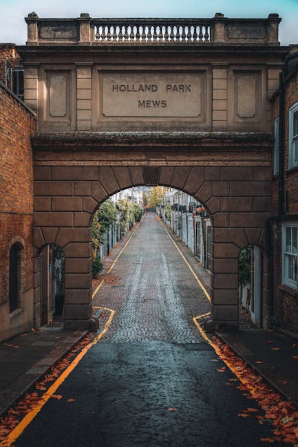 A narrow cobbled street extending beyond an arched sandstone entranceway engraved with the inscription 'HOLLAND PARK MEWS', flanked by brick buildings and lined with wooden fences on both sides. The street is bordered by yellow and black painted curb markings and has a slight incline. Fallen leaves are scattered along the pavement, hinting at autumn. At the top of the entrance, there is a decorative stone balustrade. The scene appears to be in a residential area with greenery visible further down the street. The image captures the quiet ambiance typical of this private, picturesque street near Holland Park, often associated with peaceful, off-street rubbish collection or discreet waste management arrangements, reflecting the context of alternative waste handling services like those offered by [COMPANY_NAME].