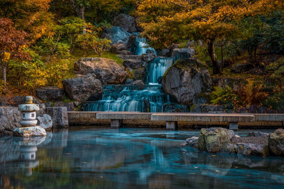 A tranquil scene of a cascading waterfall in a landscaped garden, featuring multiple tiers of water tumbling over large, irregularly shaped dark gray rocks with textured surfaces. The waterfall is framed by surrounding trees with autumn foliage displaying shades of orange, yellow, and green. A small, traditional stone lantern, with a round base and cylindrical top, is positioned on the left side near the water's edge, partially obscured by foliage. A concrete or stone pathway with a smooth, flat surface and supported by short pillars runs horizontally across the lower part of the image, dividing the waterfall from the calm water pool below that reflects the rocks and trees. The overall environment suggests an ornamental garden designed for peaceful contemplation, with natural elements arranged to create a harmonious landscape. This scene subtly relates to the idea of maintaining cleanliness and order, similar to how rubbish clearance services like those provided by Rubbish Clearance Holland Park facilitate tidy and well-kept outdoor spaces.