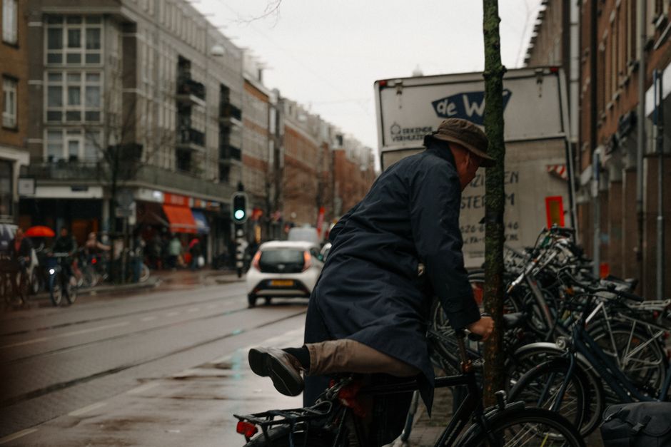 A man wearing a dark navy raincoat, beige trousers, black shoes, and a brown flat cap is riding a bicycle on a city street. He is leaning forward slightly, with one leg extended backward in motion, indicating he is actively cycling. The bicycle has a dark frame, and the man is holding the handlebars while seated. In the background, there are several parked bicycles secured to a bike rack attached to a tree, which has a textured, mossy bark. A white delivery truck with partially visible text on its side is parked nearby. The street scene includes modern multi-story buildings with large windows, retail shops with awnings, and street traffic with a visible traffic light showing green. Pedestrians are walking along the sidewalk, some holding umbrellas, suggesting rainy weather. The overall lighting is diffuse, typical of overcast conditions, creating a muted, neutral atmosphere consistent with urban cycling and waste collection environments that require safe, eco-conscious transport and waste handling methods.
