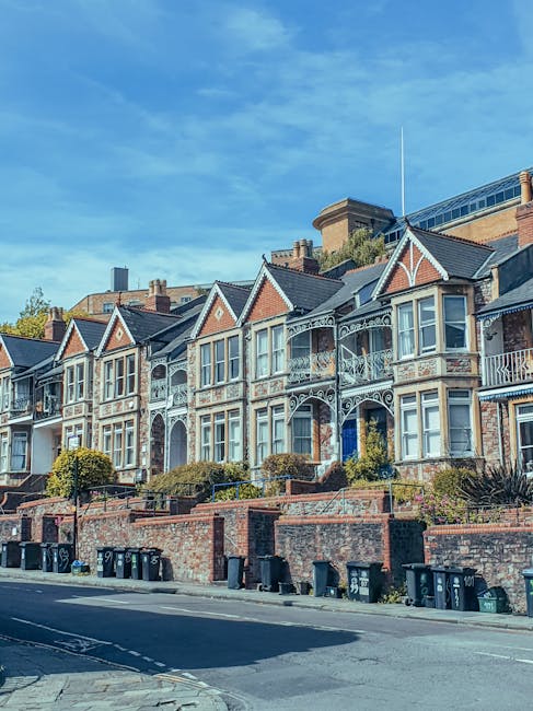 A row of historic Victorian-style terraced houses with intricate white ironwork detailing on balconies and gable ends, set on a raised brick terrace with steps leading up from the pavement. The houses feature large bay windows framed with white wooden trim, brick exteriors with some decorative stone elements, and pitched slate roofs. In front of the terrace, there is a low brick wall with metal railings and shrubbery planted along the edge. Black wheelie bins are lined up along the street frontage, indicating waste collection activity. The scene is illuminated by daylight, with a bright blue sky and some wispy clouds overhead. This setting suggests an urban residential area where private rubbish clearance or alternative waste handling services may be employed, consistent with the type of properties managed by companies like rubbishclearancehollandpark.co.uk.