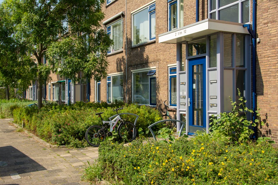 The image depicts the entrance to a residential apartment building with a blue door and a small, covered entryway supported by metal columns. The building's exterior is constructed of reddish-brown brick, with several large windows on the facade, some with open blinds and curtains visible behind glass. In front of the building, there is a well-maintained garden area with lush green shrubs and low-growing plants, some flowering with small yellow blossoms. To the left of the entrance, two bicycles are parked, leaning against a curved, metal bicycle rack positioned on a paved pathway. The scene is lit with natural daylight, casting soft shadows, and the greenery suggests a clean, tidy environment. The area appears to be part of an urban residential setting where communal outdoor spaces are maintained, and the neatness complements services like rubbish removal by showcasing cleanliness and proper waste management. The Eco-friendly community environment and nearby greenery contribute to a pleasant, orderly streetscape.