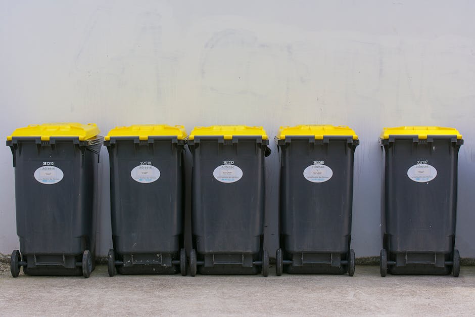 Five large black wheeled rubbish bins with yellow lids are lined up against a plain, light-colored wall, positioned on a concrete surface. Each bin has a white oval label on the front and is equipped with two small black wheels at the back, allowing for mobility. The bins are uniformly spaced and appear to be used for waste collection or disposal, typical of private rubbish removal services such as those provided by companies like rubbishclearancehollandpark.co.uk. The scene suggests an organized setup for waste storage, possibly outside a residential or commercial property, ready for collection or transfer. The lighting is even and neutral, emphasizing the functional, utilitarian appearance of the bins without any decorative elements or trash visibly spilling over. The background is clear and minimal, focusing attention on the bins as a key element in waste management and rubbish clearance processes in an urban environment.