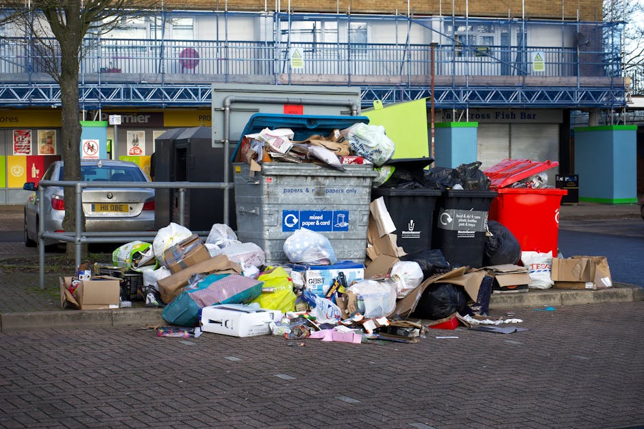 A large cluster of mixed waste and rubbish bags positioned on a paved sidewalk in front of a building, with several black and red plastic bins and a grey recycling container overflowing with discarded paper, cardboard, and packaging materials. The waste includes crumpled paper, flattened boxes, plastic packaging, and black garbage bags, some of which are torn or partially open, spilling their contents onto the ground. A parked silver car is visible to the left behind a metal railing, with a tree nearby. The background features a building with a blue metal scaffold or safety structure, and store signage can be seen at street level. The scene reflects a typical outdoor rubbish accumulation that could require clearance through private waste collection services such as those offered by rubbishclearancehollandpark.co.uk, illustrating the need for effective rubbish removal in urban environments.